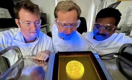 three people in safety glasses and white coats look at a glowing round yellow dish on a metal table